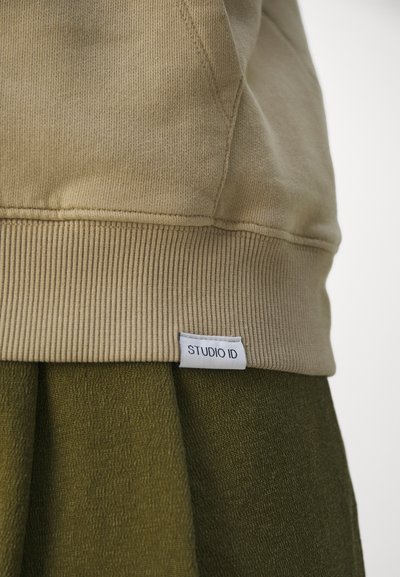 Barna pulcsi bordás szegéllyel, melyen a "STUDIO ID." feliratú fehér címke található. Texturált sötétzöld anyaggal párosítva alatta.