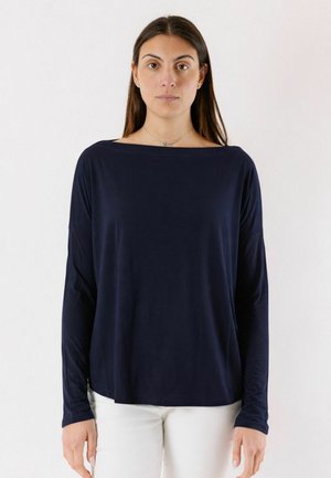 Donna in piedi su sfondo semplice, indossa una maglia blu navy a maniche lunghe con scollo a barca e pantaloni bianchi.