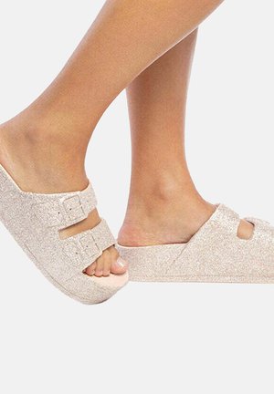 Deux pieds portant des sandales à glissière brillantes et rose clair, ornées de deux boucles, mettant en valeur le design et la texture de la chaussure.
