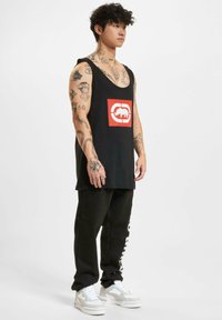 Black sleeveless tank top featuring a red graphic logo, paired with black sweatpants and white sneakers with silver accents.