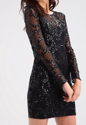 Vestido mini negro de manga larga con tela transparente y patrones florales de lentejuelas, llevado por una persona de pie con las manos entrelazadas.