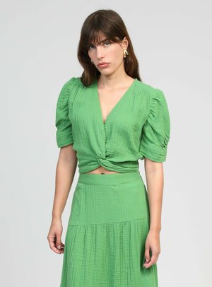 Femme aux cheveux bruns portant des boucles d'oreilles créoles dorées, un haut texturé vert à col en V avec des manches bouffantes et une jupe taille haute assortie sur un fond uni.