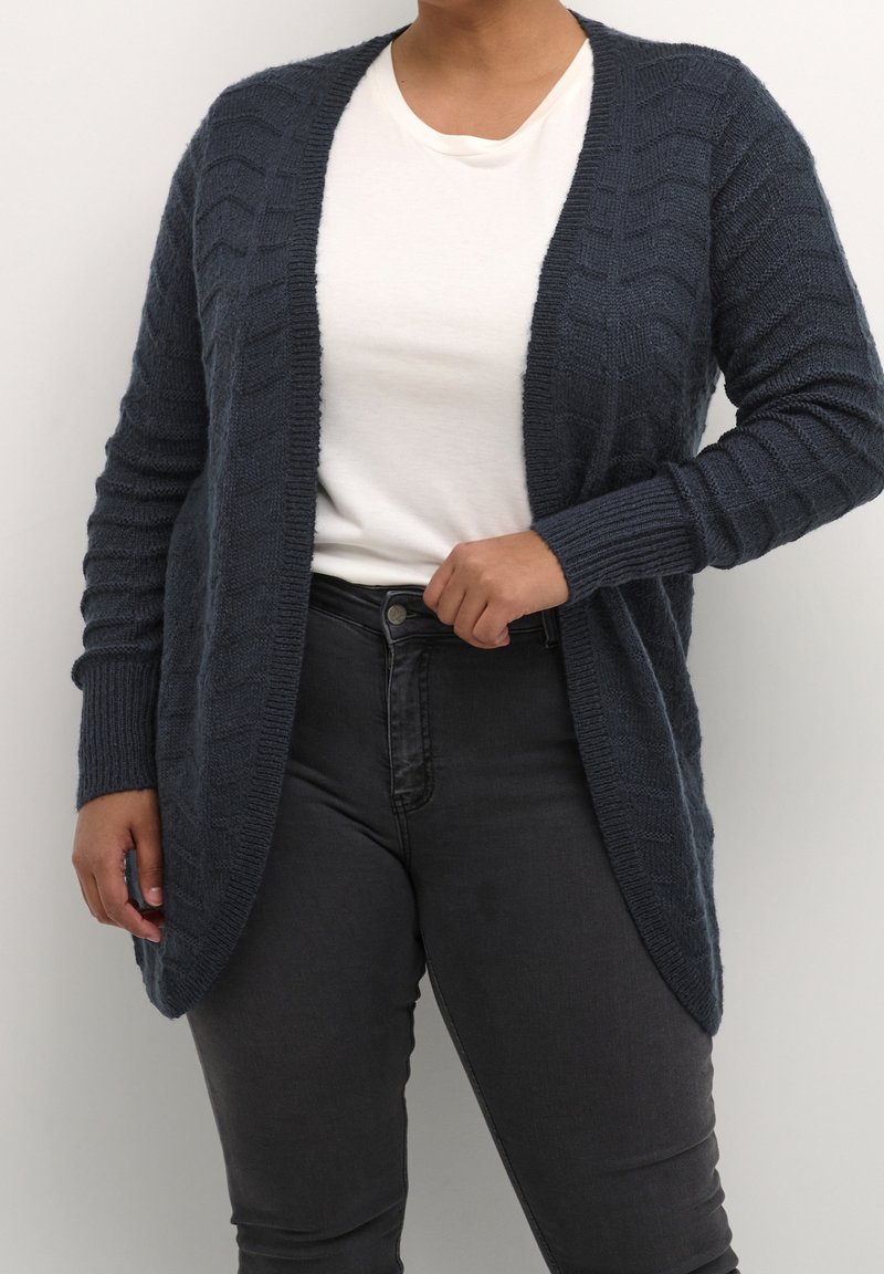 Personne portant un cardigan texturé gris foncé ouvert sur le devant, un t-shirt blanc à col rond et un jean noir ajusté, main ajustant le cardigan.