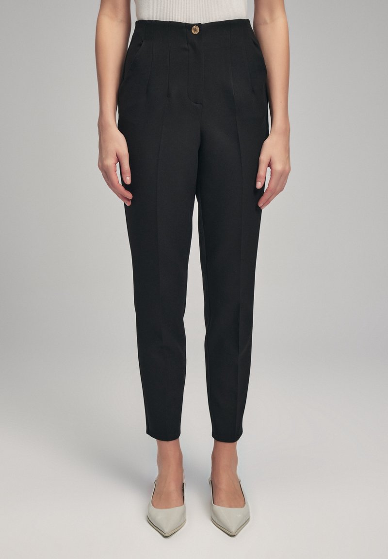 Femme portant un pantalon noir taille haute fuselé avec un bouton à l'avant et des talons blancs pointus, debout sur un fond gris clair.