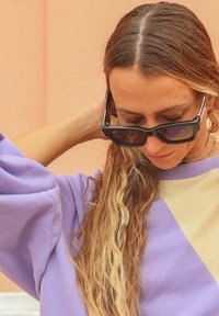 Sweat-shirt oversize violet et beige avec un motif en losanges et des poignets côtelés. Lunettes de soleil noires avec des verres sombres. Cheveux longs et ondulés.