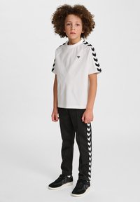 T-shirt blanc à manches courtes avec des manches à motif chevron noir, associé à un pantalon noir avec des rayures verticales blanches. Baskets noires.