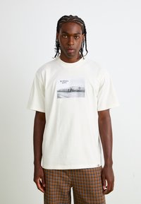Cream-colored cotton t-shirt featuring a black and white photographic print and text design on the front. Short sleeves, relaxed fit.
