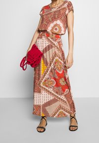 Robe maxi avec un motif en patchwork coloré dans les tons rouge, orange et crème. Elle a des manches courtes et une taille froncée. Sac en tricot rouge en complément.