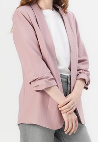 Blazer rose pâle ajusté avec une coupe décontractée, manches retroussées et une texture lisse. Porté sur un t-shirt blanc et un jean gris.