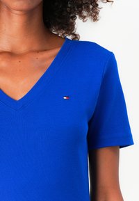 Zilā V-veida T-krekls, izgatavots no gluda kokvilnas auduma, ar īsām piedurknēm un nelielu zīmola logo sarkans, balts un zils krāsās krūšu rajonā.