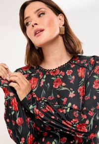 Juoda gėlių blusa su raudonomis gėlėmis, žaliomis lapelėmis, ilgomis pūstomis rankovėmis ir banguotu apykakle. Audinys yra glotnus ir švelnus.