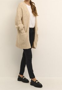 Beige Strickjacke mit geripptem Saum und Taschen, kombiniert mit engen schwarzen Jeans und schwarzen Slippern mit goldenen Akzenten.