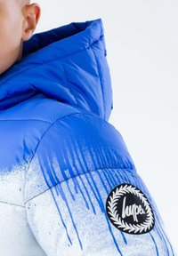 Giacca puffer blu con cappuccio, caratterizzata da un motivo a gradiente che va dal blu al bianco. Include una toppa circolare nera con logo sul maniche.