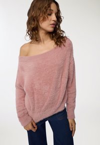 Maglione oversized rosa peloso con scollatura off-the-shoulder, polsini a coste e vestibilità rilassata, abbinato a jeans blu in denim a gamba larga.