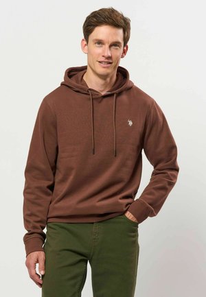 Homme portant un sweat à capuche marron et un pantalon vert, debout avec une main dans la poche, souriant légèrement, fond clair.