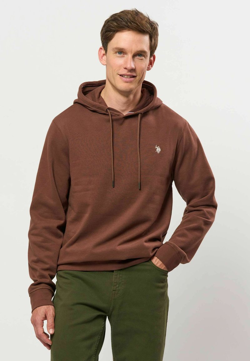 Homme portant un sweat à capuche marron et un pantalon vert, debout avec une main dans la poche, souriant légèrement, fond clair.