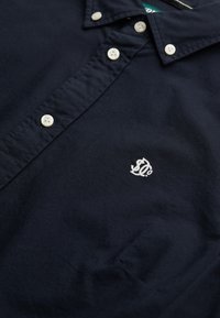 Chemise bleu marine à col avec des boutons blancs et un petit logo brodé blanc sur la poitrine.