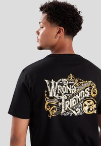 Černé bavlněné tričko s velkým zlatým a bílým grafikou "WRONG FRIENDS" na zádech, doplněné okrasnými designovými prvky.