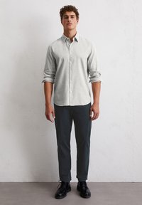 Chemise boutonnée gris clair avec des manches longues, assortie à un pantalon foncé et des chaussures noires. Tissu lisse et coupe ajustée. Design simple.