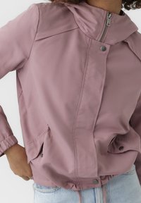 Person som bär en dammig rosa zip-up jacka med tryckknappar och elastiska ärmslut, ihop med ljusblå jeans, med handen i fickan.