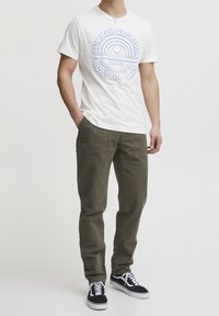 T-shirt blanc avec un motif bleu indiquant "VACANCES D'ÉTÉ", associé à un pantalon vert olive et des baskets noires à semelles blanches.