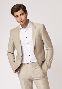 Beige angepasster Anzug mit einem weißen Hemd, ausgestattet mit braunen Knöpfen und einer subtilen Textur. Der Blazer hat eine Tasche und ein Emblem-Detail an der Revers.