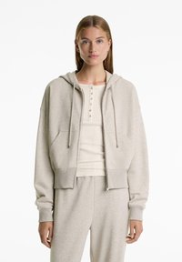 Light grey cropped hoodie in soft fabric; features a drawstring hood, front pockets, and ribbed cuffs. Paired with a matching top.