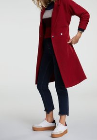 Long manteau rouge avec des boutons argentés, porté sur un pull bicolore. Jean denim foncé avec ourlets effilochés et baskets blanches à plateforme.