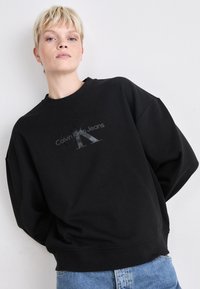 Svart oversized hettegenser laget av mykt stoff, med en fremtredende grå Calvin Klein Jeans-logo på forsiden. Koselig og avslappet design.