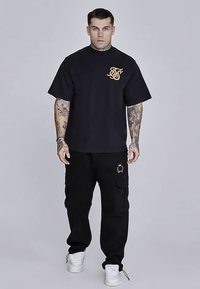 T-shirt noir oversize avec logo doré sur la poitrine, associée à un pantalon cargo noir avec poches latérales et accents de quincaillerie dorée. Baskets blanches.