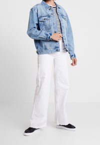 Person som bär en ljusblå denimjacka över en svart-vit randig skjorta, vita vida byxor och svarta sneakers med vita sulor.