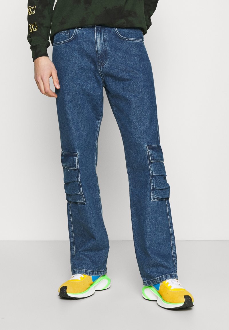 Blue denim cargo pants with side pockets, straight leg design, and faded details worn with yellow and green sneakers.