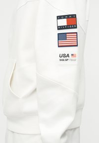 Bílý rukáv s třemi nášivkami: logo Tommy Hilfiger, americká vlajka a text "USA Sail GP Team" s malou vlajkou.