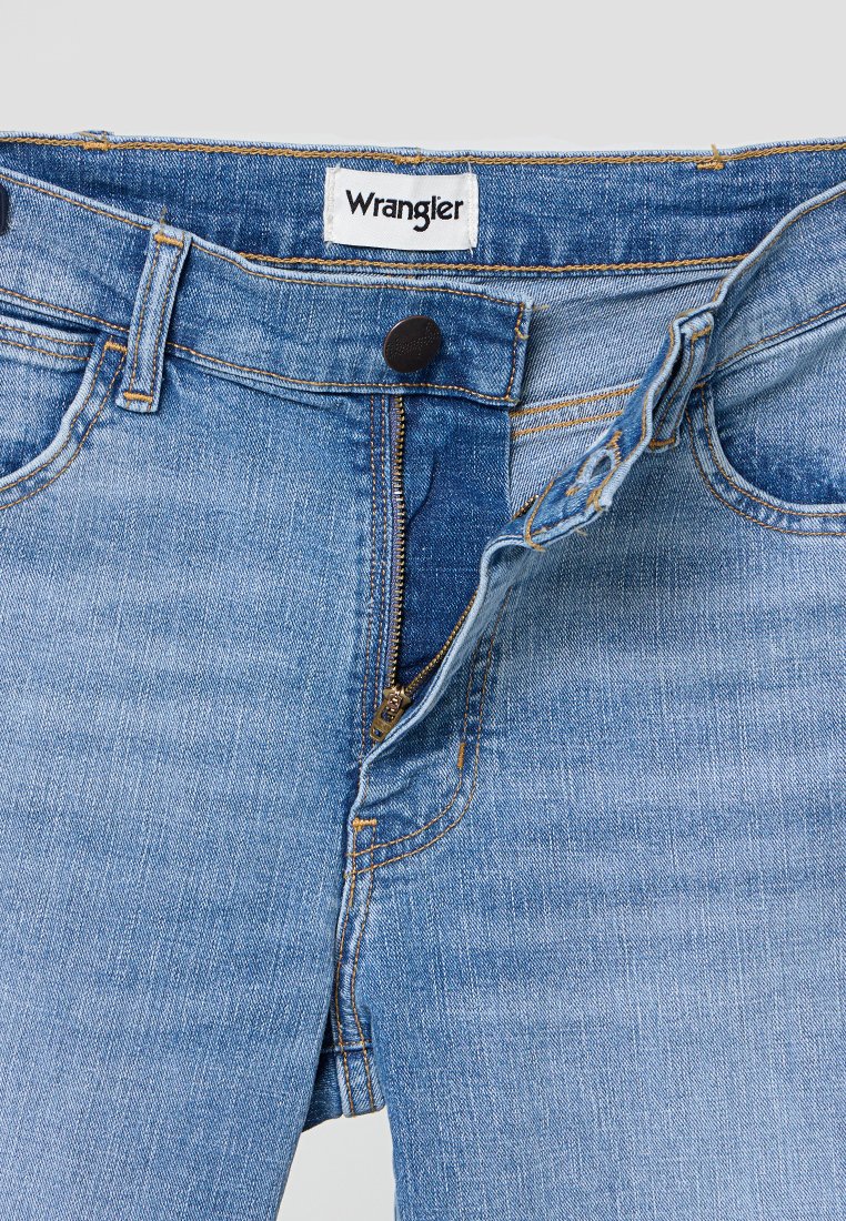 Jeans z denimu w jasnoniebieskim kolorze, z zamkiem błyskawicznym i zapięciem na guzik. Widoczna etykieta marki Wrangler przy pasie.