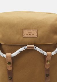 Sac à dos en toile marron avec rabat supérieur, accents en cuir et fermeture en corde tissée ornée de détails noirs et blancs. Logo "Doughnut" visible.