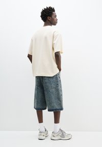 Beige oversize tričko, volné modré denimové šortky a šedé tenisky s reflexními detaily, zobrazené zezadu na bílém pozadí.