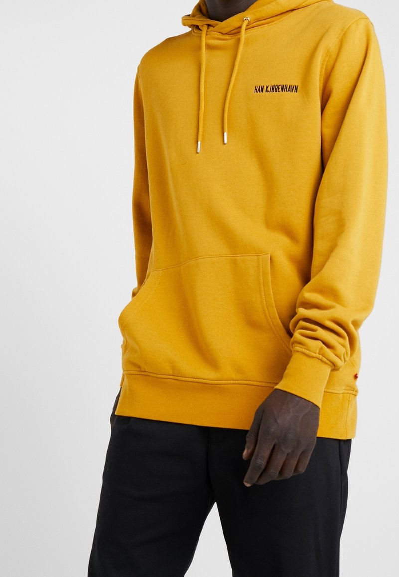 Personne portant un sweat à capuche jaune moutarde avec poche avant et pantalon noir, texte « HAN KJØBENHAVN » sur la poitrine.