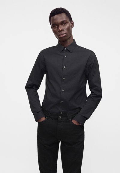 Uomo che indossa una camicia nera aderente con bottoni e pantaloni neri, in piedi con le mani nelle tasche davanti a uno sfondo chiaro e semplice.