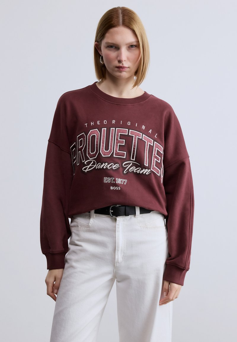 Μαρόν φούτερ με φαρδιά εφαρμογή και μανσέτες, με το κείμενο "PROUETTE Dance Team" σε λευκό και χρυσές λεπτομέρειες. Συνδυάζεται με λευκό παντελόνι και μαύρη ζώνη.