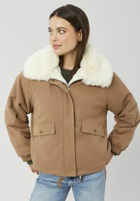 Veste rembourrée marron avec un grand col en fausse fourrure crème, fermeture éclair frontale et deux poches basses. Comprend des poignets élastiques et un ourlet à cordon de serrage.