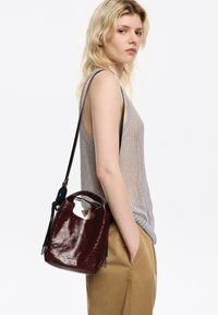 Bolso de mano de charol color burdeos con apertura redondeada, correa delgada para el hombro y dos adornos con borlas. Lo lleva una persona con una prenda superior clara.