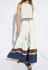 Femme portant un haut sans manches blanc et une longue jupe à volants avec un motif de tie-dye bleu et marron, associée à des sandales noires.