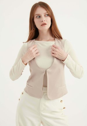 Beige getailleerde vest met een ronde halslijn, zijkragen en een gladde textuur. Het model draagt eronder een licht geribbeld topje.