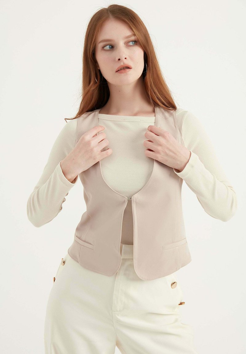 Beige, taillierter Weste mit Rundhalsausschnitt, Seitentaschen und glatter Textur. Das Model trägt ein leicht geripptes Oberteil darunter.
