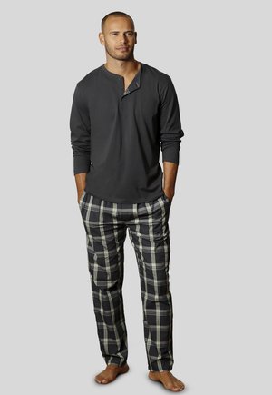 Man standing barefoot wearing a dark long-sleeve henley shirt and black and white plaid pajama pants against a plain gray background.