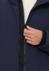 Giacca blu navy con cerniera nera testurizzata e fodera in rete, con un primo piano di una mano che la chiude con la cerniera, mettendo in mostra il suo design e materiale.