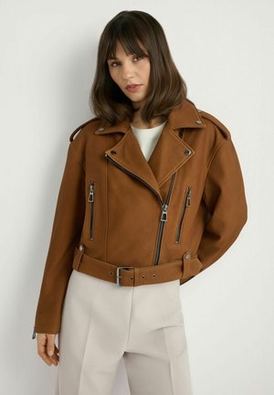 Kobieta w brązowej skórzanej kurtce motocyklowej z zamkami błyskawicznymi i paskiem, w połączeniu z jasnobeżowymi spodniami na jednolitym tle.