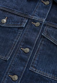 Denimjakke i mørkeblåt stof med metalliske knaplukninger og kontrasterende beige syninger; inkluderer brystlommer med klap.