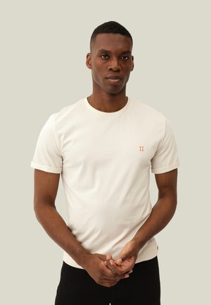 Homme aux cheveux courts portant un t-shirt blanc ajusté et un pantalon noir, debout devant un fond clair uni, les mains posées légèrement jointes.