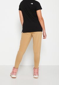 Osoba v černém tričku The North Face, béžových legínách a růžových zimních botách, stojící na bílé pozadí čelem odvrácená.